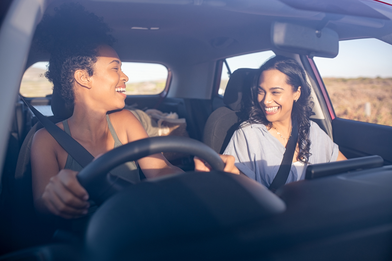 Young beautiful friends laughing while traveling in car during a road trip. Two multiethnic women driving in car and having fun. Two attractive girls talking together while driving in a car through the countryside on a sunset.