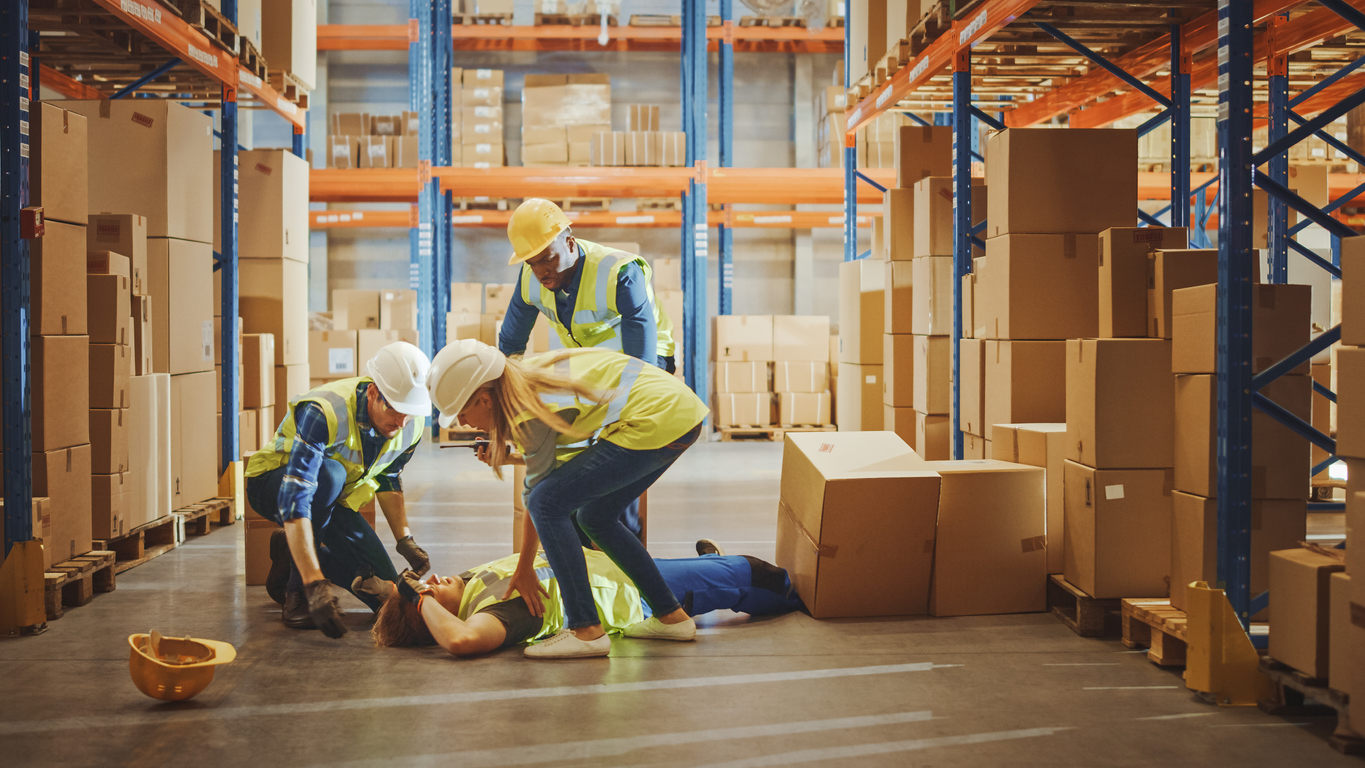 Warehouse Worker Has Work Related Accident Falls while Trying to Pick Up Cardboard Box from the Shelf. Colleagues Call for Help and Medical Assistance. Injury at Work.