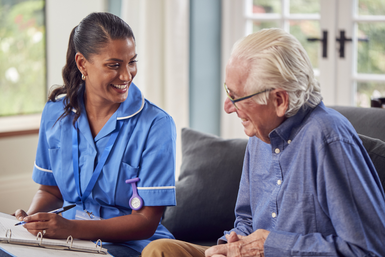 Senior Man At Home Talking To Female Nurse Or Care Worker In Uniform Making Notes In Folder