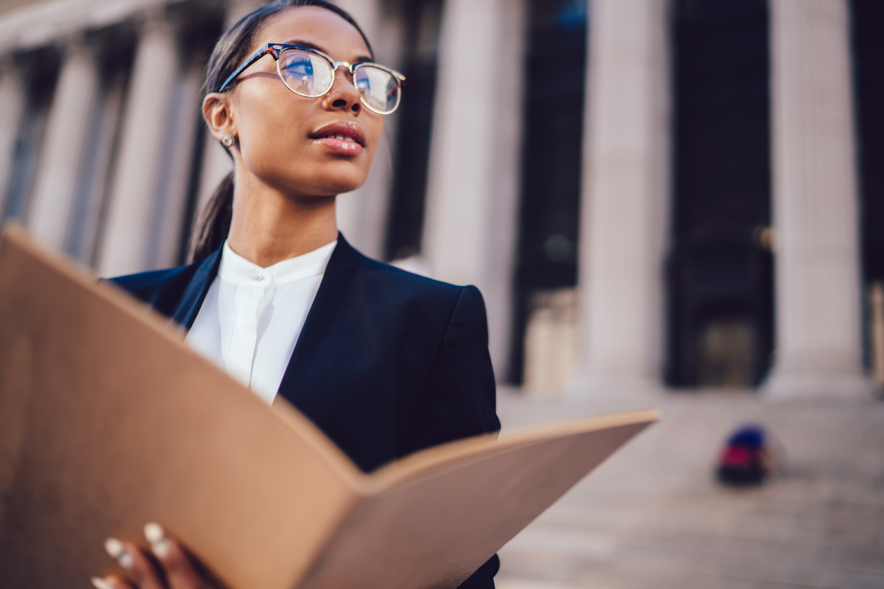 Thoughtful African American businesswoman in optical spectacles holding documents looking away while standing in urban setting with copy space. Female dark skinned student of high economic university