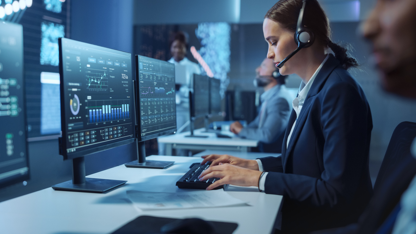 Happy Beautiful Technical Customer Support Specialist Having a Headset Call while Working on a Computer in a Dark Monitoring and Control Room Filled with Colleagues and Display Screens.