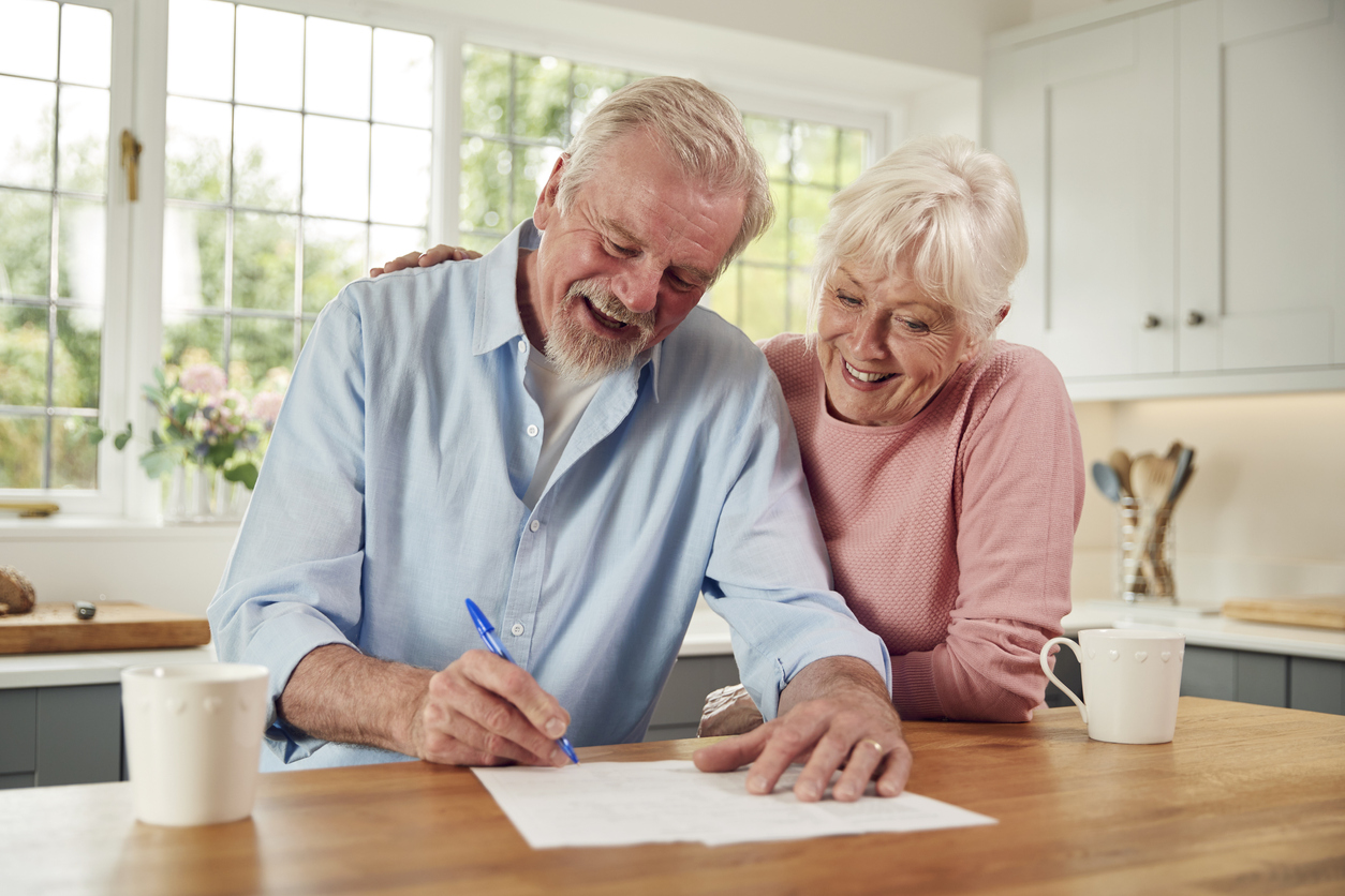 Retired Senior Couple Sitting In Kitchen At Home Signing Financial Document