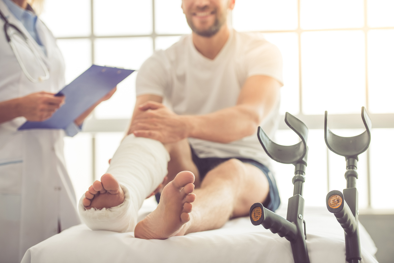 Cropped image of beautiful female medical doctor listening to handsome patient with broken leg and making notes while working in her office