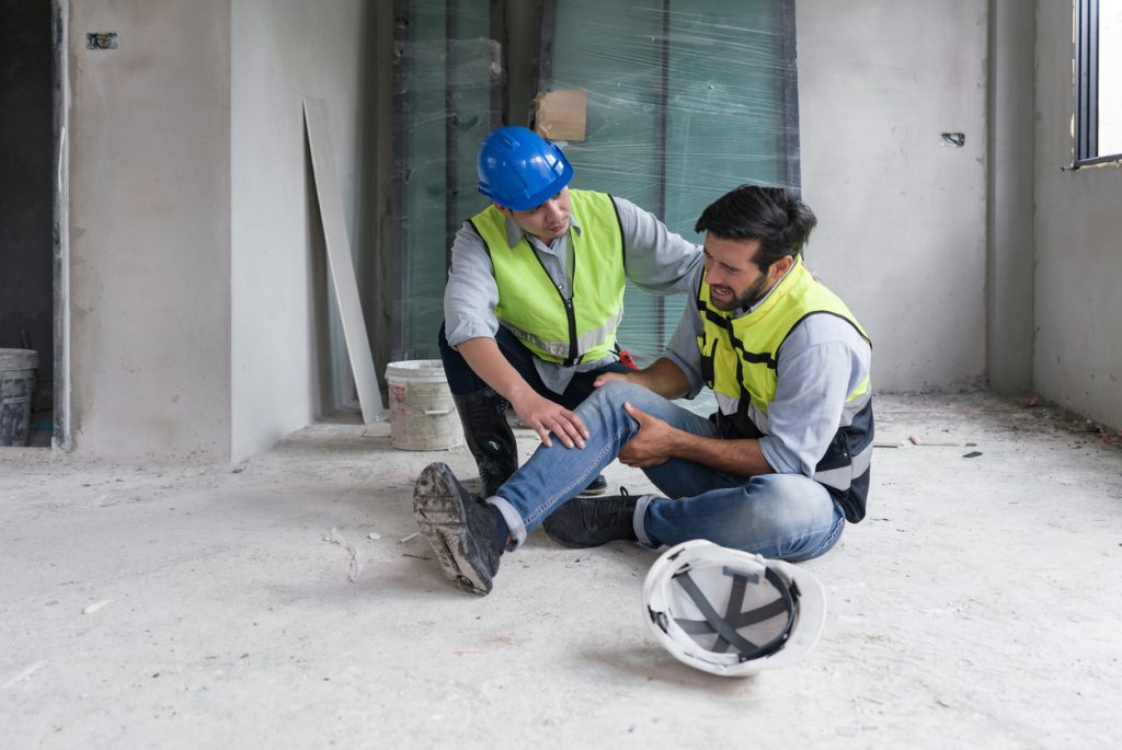 young caucasian man in safety vest gripped his knee and screams in pain. due to injuries at the construction site. his asian friend comes and help him to stand.