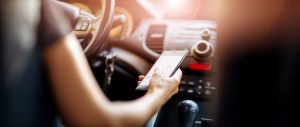 Driver driving a car on asphalt road in summer day at park. Woman at steering wheel with smartphone in her hand.