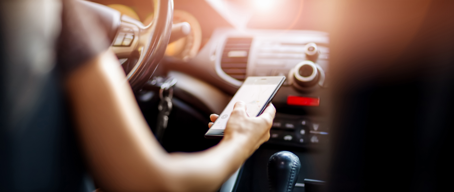 Driver driving a car on asphalt road in summer day at park. Woman at steering wheel with smartphone in her hand.