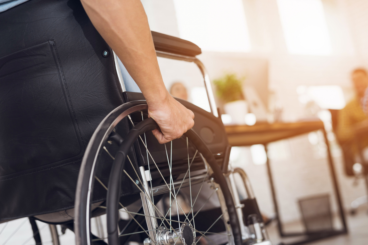 A disabled man is sitting in a wheelchair. He holds his hands on the wheel. Nearby are his colleagues