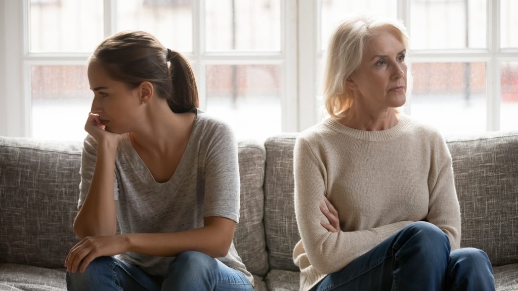 elderly mother and grown up daughter sit on couch separately