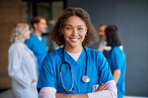 Confident happy woman nurse smiling with crossed arms with healthcare team in background. Multiethnic medical professional in scrub ready to assist patients. Portrait of reliable confident nurse standing proudly, surrounded by colleagues in a private clinic.